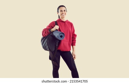 Studio shot of smiling female athlete in workout outfit with gym mat and sports bag. Happy beautiful woman wearing comfortable red hoodie and black leggings standing isolated on light beige background - Powered by Shutterstock