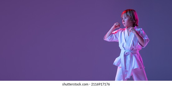 Studio Shot Of Sportive Kid, Male Taekwondo, Karate Athletes In Doboks Posing Isolated On Purple Background In Neon. Concept Of Sport, Education, Skills, Martial Arts, Healthy Lifestyle.