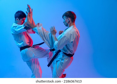 Studio shot of sports training of two karatedo fighters in doboks isolated on blue background in neon. Concept of combat sport, challenges, skills. Sportsmen practicing base technique - Powered by Shutterstock
