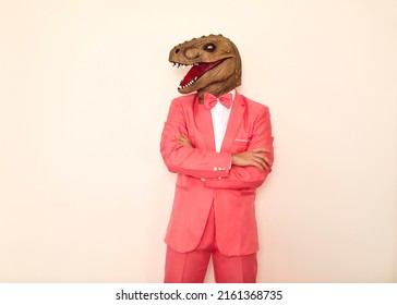 Studio shot of strange guy wearing wacky ugly dinosaur mask. Man in pink party suit, bow tie and funny silly scary masquerade lizard mask standing with his arms folded isolated on white background