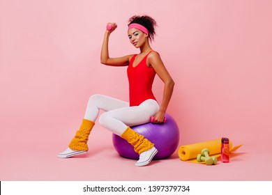 Studio Shot Of Strong Girl Posing With Sport Equipment. Shapely African Lady Sitting On Fitness Ball.