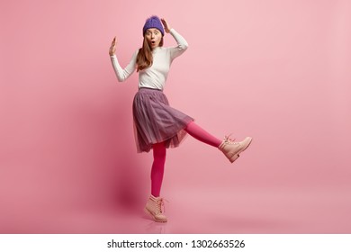 Studio shot of stupefied emotional frighted female model stands sideways, wears blue warm headgear, skirt, bright pink tights, boots, raises legs, overwhelmed by unexpected news. Omg concept