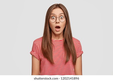 Studio shot of stupefied emotive woman keeps jaw dropped, reacts on sudden news from friend, wears round big glasses, isolated over white background. Astonished bugged eyed girlfriend stares indoor