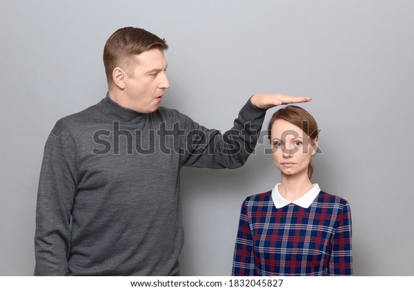 Studio shot of surprised amazed tall man showing\
height of short woman, both are wearing casual clothes, standing\
over gray background. Concept of diversity of people\'s heights,\
tall and short persons