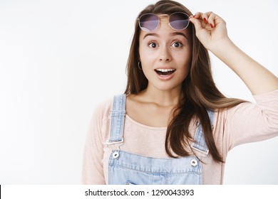 Studio shot of surprised attractive young woman unexpected to see friend in crowd taking off sunglasses raising eyebrows and open mouth in smile from amazement reacting to awesome surprise