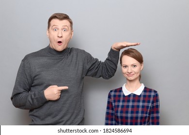 Studio Shot Of Surprised Tall Man Showing Height Of Short Woman, Pointing At Her With Finger, Both Are Standing Over Gray Background. Concept Of Diversity Of People's Heights, Tall And Short Persons