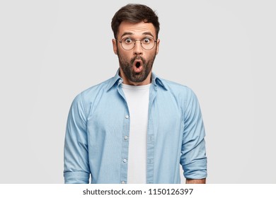 Studio Shot Of Terrified Unshaven Man Stares With Eyes Full Of Disbelief, Being Surprised By Curious Revelation, Wears Blue Shirt, Isolated Over White Studio Wall. People, Reaction, Facial Expressions