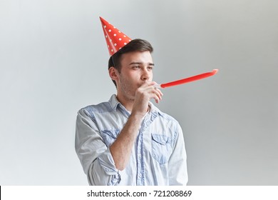 Studio Shot Of Unshaven Young Caucasian Male Blowing Whistle While Celebrating His Friend's Birthday, Having Relaxed And Cheerful Expression On His Face. Happy Guy Posing With Red Party Horn