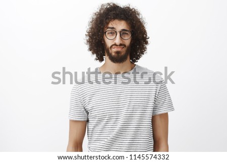 Studio shot of unsure good-looking bearded male with curly hair in trendy black glasses, lifting eyebrown and looking with doubt at camera, being displeased over gray background