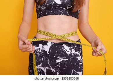 Studio Shot Of Woman Measure Her Waist Belly By Metre Stick, Slim Model Posing Isolated Over Yellow Studio Background, Faceless Female Wearing Black Top And Leggins. Sport And Fitness Concept,