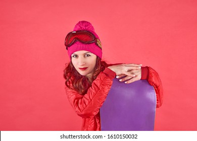 Studio Shot Woman In Rad Wearing Ski Clothes Standing In Red Background With Purple Snowboard And Ski Mask