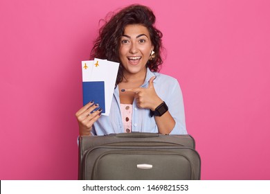 Studio Shot Of Woman With Suitcase, Showing Her Tickets For Plane In Her Hands With Fore Finger, Isolated Over Rosy Background, Model With Toothy Smile, Experesses Happyness, Wearinf Stylish Outfit.