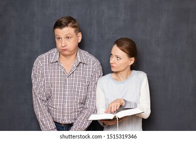 Studio Shot Of Woman Trying To Pay Attention Of Displeased Man To Text In Book While He Is Bored And Tired Of Reading And Studying, Being Not Interested In Tedious Activity, Over Gray Background