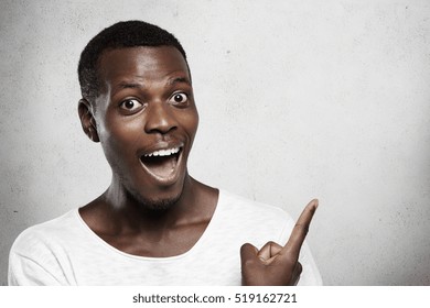 Studio shot of young African man dressed in white t-shirt opening his mouth widely in surprise, looking shocked with big sale prices, showing something at blank wall with his finger. Body language
