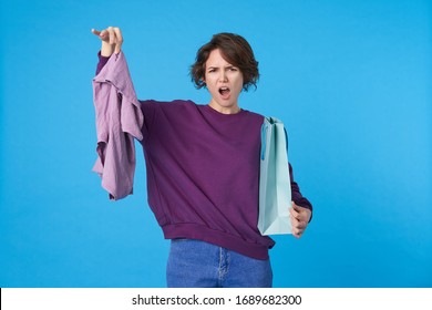 Studio shot of young annoyed dark haired curly lady keeping her hand raised with purple clothes while standing over blue background, don't like her present