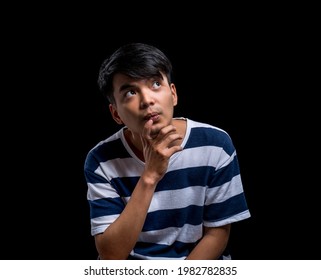 Studio Shot Of Young Asian Man Having Doubts And With A Confused Face, Hand On Chin Thinking About The Question, Man Looking Up, Solution Of A Problem, Makes Up An Idea. Isolated Black Background