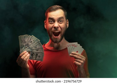 Studio Shot Of Young Excited Man Posing With A Stack Of Playing Cards And Ward Of Dollar Banknotes In Hands Over Black Background With Smoke Effect. Gambling, Sport Betting, Poker, Casino Concept.