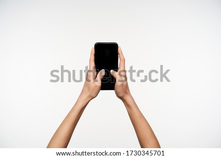 Studio shot of young female's raised hands keeping mobile phone while typing text with thumbs, being isolated over white background. Hand gesturing and signs concept