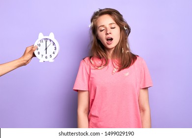 Studio Shot Of Young Good Looking Female In Pink Pyjamas Yawning, Trying To Wake Up, Hates Early Mornings, Had Bad Sleep, Disturbing Dreams, Posing Over Light Purple Background.