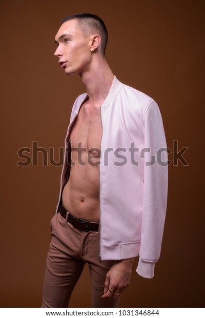 Studio shot of young\
handsome androgynous man wearing pink jacket fully unbuttoned\
against brown background