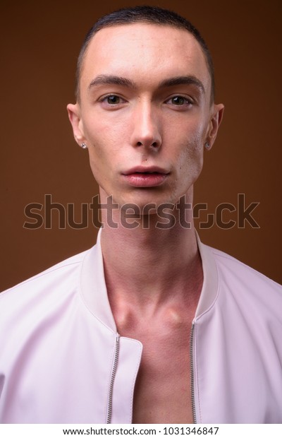 Studio shot of young\
handsome androgynous man wearing pink jacket fully unbuttoned\
against brown background