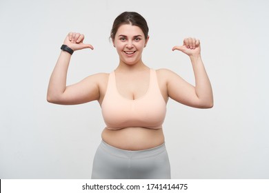 Studio Shot Of Young Happy Cute Dark Haired Fatty Lady With Ponytail Hairstyle Thumbing On Herself While Looking Cheerfully At Camera, Standing Over White Background