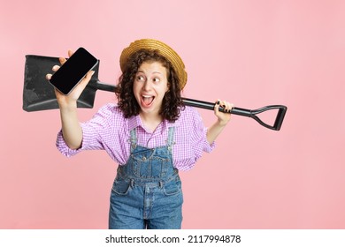 Studio Shot Of Young Happy Girl, Female Gardener In Work Uniform And Hat Using Phone Isolated On Pink Background. Concept Of Job, Emotions, Agronomy. Funny Meme Emotions. Copy Space For Ad