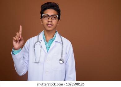 Studio shot of young Indian boy doctor wearing eyeglasses against brown background
