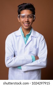 Studio shot of young Indian boy doctor wearing protective glasses against brown background