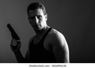 Studio Shot Of Young Muscular Persian Man Holding Gun While Thinking And Looking Back In Dark Room