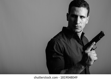 Studio Shot Of Young Muscular Persian Man Holding Gun While Thinking And Looking Back Ready For Action Against Gray Background