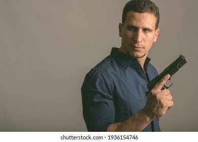 Studio Shot Of Young Muscular Persian Man Holding Gun While Thinking And Looking Back Ready For Action Against Gray Background