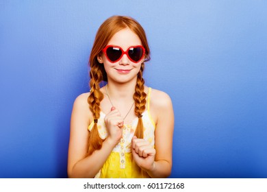 Studio shot of young preteen 9-10 year old redhead girl wearing heart shape sunglasses, standing against blue purple background