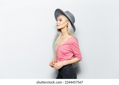Studio side portrait of young attractive blonde girl with wireless earphones in ears, wearing grey hat and pink shirt, on white background.