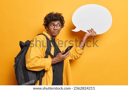 Studio sideway photo of young dissatisfied Hindu male student standing on yellow background holding white speech bubble with blank space for promotion and smartphone got message with bad test result