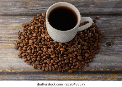 Studio top-down angle photograph capturing a white textured ceramic cup filled with aromatic black coffee, positioned over a rich composition of scattered roasted coffee beans on a rustic wooden table.