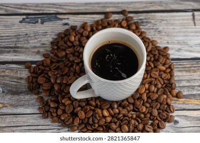 Studio top-down angle photograph capturing a white textured ceramic cup filled with hot black coffee, positioned over a rich composition of scattered roasted coffee beans on a rustic wooden table.