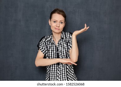 Studio Waist-up Portrait Of Bewildered Caucasian Young Woman Wearing Checkered Shirt, Looking With Goofy Expression At Camera, Raising Hand, Being Puzzled Because Of Happened, Over Gray Background