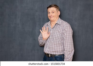 Studio waist-up portrait of cheerful friendly man dressed in casual clothes, waving his hand like saying goodbye, smiling kindly, looking with silly funny expression at camera, over gray background