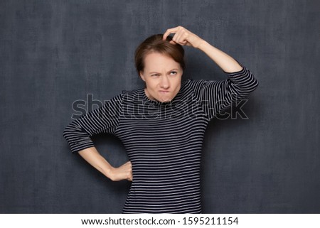 Studio waist-up portrait of funny thoughtful focused puzzled girl wearing jumper, scratching head with finger, intently thinking, planning or making difficult choice, isolated over gray background
