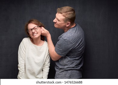 Studio Waist-up Shot Of Dissatisfied Man Frowning Face, Pulling Woman By Ear While Scolding, Woman Is Grimacing From Pain, Over Gray Background. Strict Husband And Misbehaving Wife