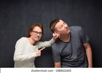 Studio Waist-up Shot Of Enraged Woman Frowning Face, Pulling Man By Ear And Pointing At Him While Scolding, Man Is Wincing From Pain, Over Gray Background. Strict Wife And Misbehaving Husband