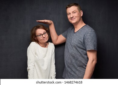 Studio Waist-up Shot Of Tall Man Smiling And Showing With Hand At Height Of Short Girl Standing Beside Him And Looking With Perplexity At Camera, Over Gray Background. Variety Of Person's Heights