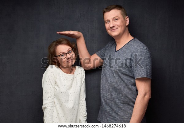 Studio waist-up shot of tall man smiling and\
showing with hand at height of short girl standing beside him and\
looking with perplexity at camera, over gray background. Variety of\
person\'s heights
