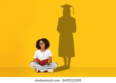 A studious young girl sits cross-legged reading a book, with her shadow on a yellow background illustrating a graduate in cap and gown, symbolizing future academic success - Powered by Shutterstock