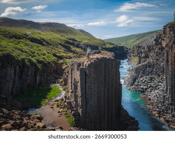 Studlagil Canyon in Iceland features dramatic basalt columns, a turquoise river, lush green hills, and a person standing atop the formations.