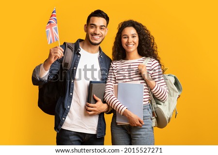 Study Abroad. Cheerful Young Arab Man And Woman With Backpacks Holding British Flag, Portrait Of Happy Smiling Students With Union Jack And Workbooks Over Yellow Studio Background, Copy Space