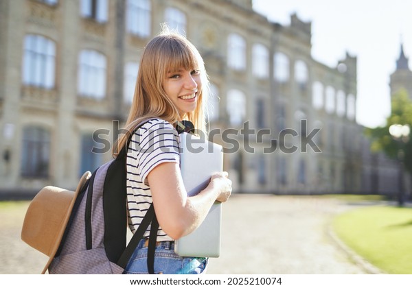 Study abroad, high school study or educational
concept. Cute happy smiling blonde college or university student
girl with backpack, hat and laptop in campus at summer. High
quality image