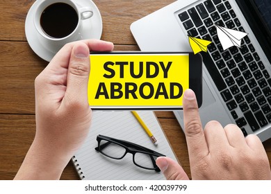 STUDY ABROAD  message on hand holding to touch a phone, top view, table computer coffee and book