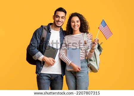 Study Abroad. Smiling Middle Eastern Students Couple Posing With American Flag In Hand, Happy Young Arab Man And Woman With Backpacks And Workbooks Standing Over Yellow Studio Background, Copy Space